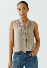 Ärmellose beige Bluse mit einer Schleife am Kragen, aus weichem Stoff und leichtem Material, kombiniert mit blauen High-Waist-Jeans.