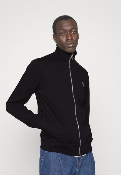 Sweat-shirt à zip noir avec un col haut, featuring une fermeture éclair argentée et des poignets côtelés, porté sur un pantalon en denim bleu.