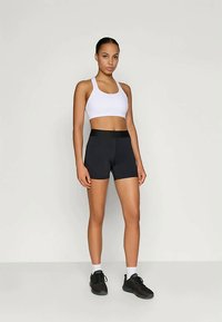 Čierne športové šortky s vysokým pásom, skombinované s bielou športovou podprsenkou. Modelka stojí na neutrálne pozadí a predvádza tento outfit.