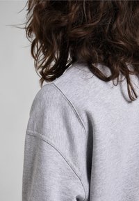 Capelli castano scuro e ricci che riposano sulla spalla di una persona che indossa un felpa grigia chiara su uno sfondo grigio uniforme.