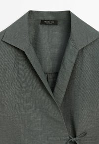 Chemise en lin gris avec un grand col, un design croisé et un accent noué à la taille. La texture est légèrement tissée, offrant une apparence douce.