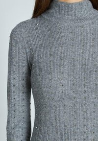 Maglione grigio a collo alto con coste, caratterizzato da borchie metalliche disposte in un motivo a pois su tutta la superficie, che mette in risalto la texture e il design.