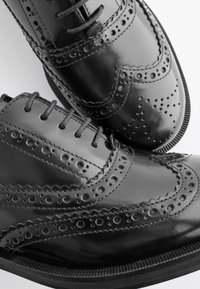 Scarpe brogue in pelle nera con dettagli traforati, design a punta alata e punta arrotondata. Visibili lacci e suola esterna con texture a contrasto.