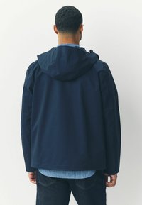 Chaqueta con capucha en color marino, confeccionada en material tejido suave, con un corte relajado y un dobladillo curvado. Llevada sobre una camisa a rayas azul claro.