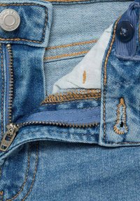 Jeans i denim med ljusblå färg, utrustade med metallblixtlås och bronsfärgad söm. Insidan avslöjar ett subtilt mönster på tyget.