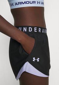 Černé sportovní kraťasy s kontrastní levandulovou vnitřní vrstvou, s širokým černým pasem a nápisem "UNDER ARMOUR" a bočními kapsami.