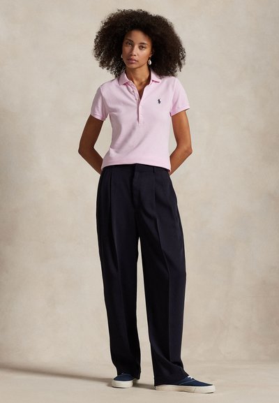 Femeie cu păr buclat, îmbrăcată într-un tricou polo roz deschis, pantaloni negri largi și adidași albastre, stând pe un fundal neutru.