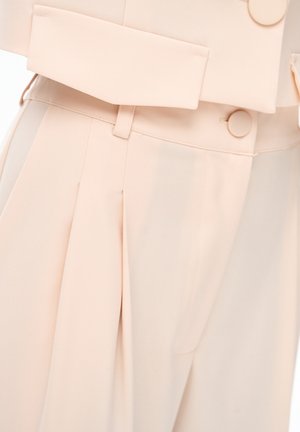 High-waist beige trousers with front pleats, matching fabric-covered buttons, belt loops, and a flap pocket detail.