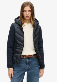 Chaqueta acolchada de puffer en color marino con capucha y mangas de punto en contraste. Frente con cremallera, bolsillos laterales, llevada sobre un suéter crema y jeans de cintura alta.