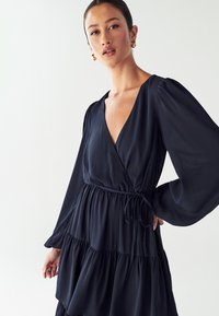 Temno mornarsko obleko z zavihki in dolgimi napihnjenimi rokavi, globokim V-izrezom, pasom na vrvico in večplastnim robom z obliki volanov. Gladek občutek, lahek material.