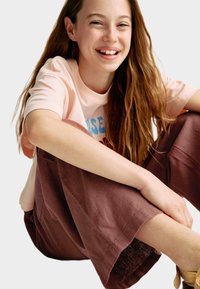 Lichtroze T-shirt met blauw grafisch ontwerp, gecombineerd met losse, donkerbruine, gestructureerde cropped pants. Model zit met een ontspannen houding.