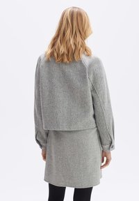 Femme blonde portant une veste longue à manches gris texturé et une jupe assortie, se tenant de dos face à un fond uni.