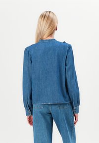 Denim top in light blue with long sleeves, gathered shoulder detail, and a rounded neckline. Features buttoned cuffs and a straight cut.
