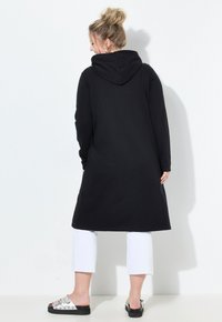 Woman viewed from behind wearing a long black hooded coat, white cropped pants, and silver sandals with black soles.