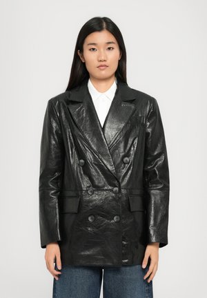 Blazer double boutonnage en cuir noir avec une texture brillante, revers crantés, deux poches avant et six boutons noirs, porté sur une chemise blanche.