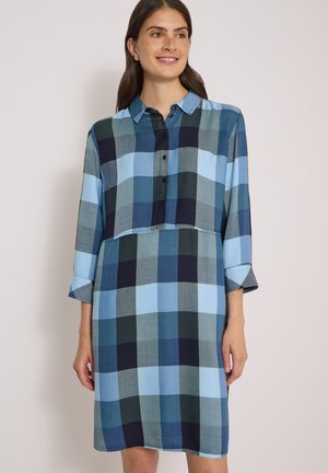 Femme portant une robe-chemise à manches longues à carreaux bleus et verts avec col et boutons, souriant légèrement vers la droite.