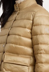 Beige-täytteinen takki, jossa vaakasuora tikattu kuvio, korkea kaulus, hopeiset yksityiskohdat ja edessä tasku, jossa on kirjailtu logo.