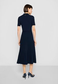 Temno modra rebrasta obleka z oprijetim zgornjim delom in naborki na krilu, z kratkim ovratnikom. Kombinirana s temno modrimi čevlji z visokimi petami.