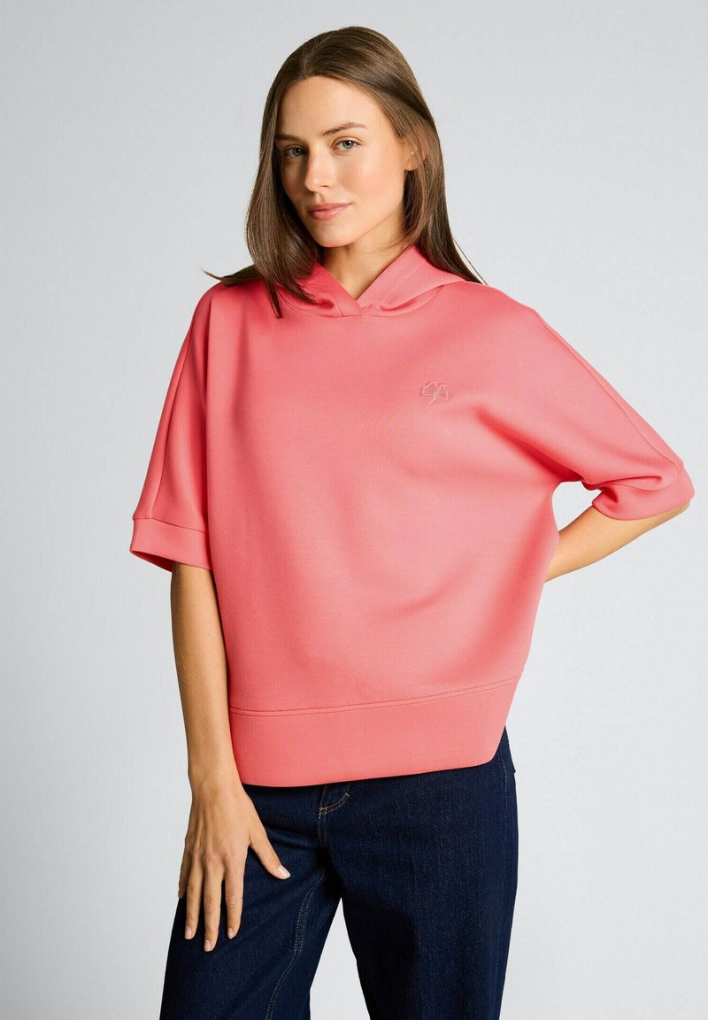 Jonge vrouw die een losse koralen korte mouwen hoodie en donkerblauwe spijkerbroek draagt, poserend met één hand in de zij tegen een effen achtergrond.