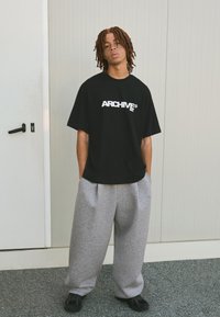 Černé oversized tričko s bílým logem "ARCHIVE", v kombinaci se šedými tepláky a černými botami. Hladký materiál; volný střih.