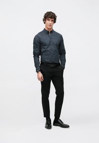 Μπλε πουκάμισο με κουμπιά και γεωμετρικό σχέδιο, σε συνδυασμό με μαύρο παντελόνι στενής εφαρμογής και μαύρα δερμάτινα παπούτσια. Το μοντέλο στέκεται όρθιο.