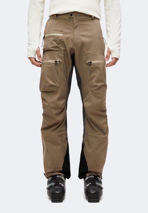 Personne portant un pantalon cargo marron avec plusieurs poches zippées et des bottes de ski noires, debout devant un fond uni.