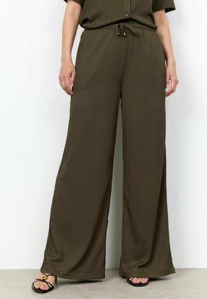 Personne portant un pantalon large vert olive avec une taille à cordon et des sandales noires à brides, debout sur un sol gris.