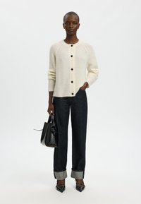 Krém színű kardigán kerek nyakkal, gombos lezárással és bordázott textúrával; sötét farmernadrággal, felfordított szárakkal a bokáknál és fekete magassarkúval párosítva.
