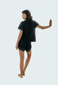 Czarny zestaw piżamowy z bawełny, składający się z krótkiego topu z białymi wykończeniami i pasujących szortów. Modelka stoi boso, odwrócona plecami.