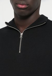 Artimas planas asmens, nešiojančio juodą megztinį su sidabro spalvos užtrauktuku, kuris yra dalinai atsegtas prie apykaklės, atidengiantis kaklą ir lūpas.