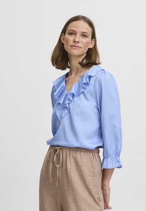 Blusa azzurra chiara con collo a volant e maniche a tre quarti, abbinata a pantaloni beige con coulisse. Tessuto morbido, vestibilità rilassata.