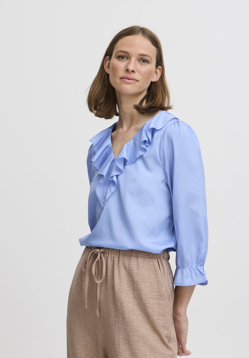 Blusa azzurra chiara con collo a volant e maniche a tre quarti, abbinata a pantaloni beige con coulisse. Tessuto morbido, vestibilità rilassata.