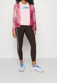 Růžovo-červená tie-dye lehká bunda, růžové tričko s logem, hnědé legíny s bílými bočnicemi a bílé sportovní boty s modrými tkaničkami.