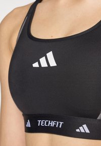 Black sports bra made of stretchy material featuring a white triangular logo. Wide black band with "TECHFIT" printed in white.