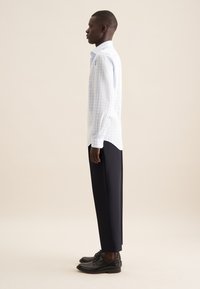 Camicia a maniche lunghe a quadri blu chiaro con orlo curvo, indossata con pantaloni scuri su misura e scarpe nere, vista di profilo.