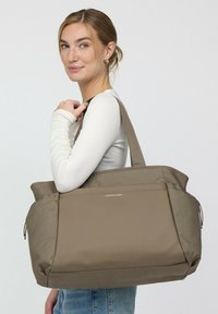 Vrouw met blond haar in een wit shirt met lange mouwen, draagt een grote taupe draagtas over haar schouder, zijwaarts gekeerd, met een lichte glimlach.