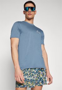 Blue short-sleeve t-shirt with a rounded neckline and a white logo on the left chest, paired with multi-colored patterned shorts.