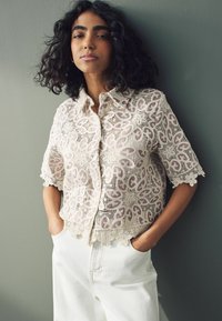 Bluse i blonde i creme med lyserøde paisley mønstre, korte ærmer, knapfront og scalloped kanter, parret med hvide højtaljede bukser.