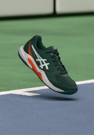 Must ja oranž spordijalats valge logoga, paelte ja paksu tallaga sinise ja rohelise tennisekatte pinnal.