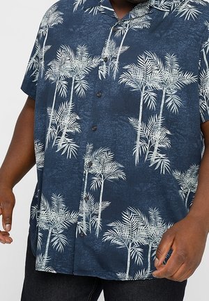 Zbliżenie osoby noszącej granatową koszulę na krótki rękaw z białymi nadrukami palm.