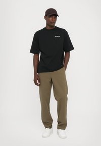 Tricou negru cu mânecă scurtă și logo alb, asortat cu pantaloni de culoare verde măsliniu și adidași albi. Modelul poartă o șapcă maro.