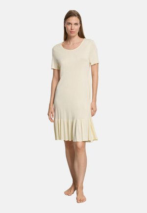 Vrouw staat op blote voeten en draagt een crèmekleurige jurk met korte mouwen en een knielange rok met ruches aan de zoom, tegen een effen witte achtergrond.