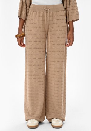 Beżowe dzianinowe spodnie o szerokich nogawkach z regulowanym sznurkiem w talii, noszone z pasującym kardiganem, drewnianą bransoletką i białymi trampkami na modelce.