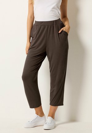 Mujer vistiendo pantalones cortos sueltos de color marrón oscuro con bolsillos, una camiseta sin mangas blanca y zapatillas blancas sobre un fondo liso.