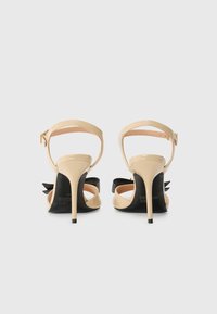 Sandale cu toc înalt din piele patent bej, cu accent de fundă neagră, brâu pentru gleznă și toc stiletto, având o textură netedă și un design elegant.