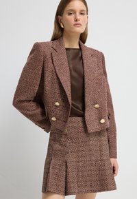 Conjunto de chaqueta de doble botonadura y falda en una tela texturizada color burdeos con botones dorados y un sutil patrón de cuadros. Top marrón suave debajo.