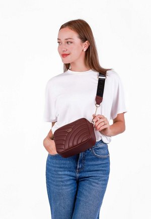Hnědá crossbody taška s prošívanou texturou, zlatým logem a popruhem s kontrastním černým písmem. Hladký materiál, obdélníkový tvar.