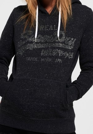 Donna che indossa un felpa nera a pois con lacci bianchi e una scritta lucida "Superdry" sul fronte, mani nella tasca anteriore.