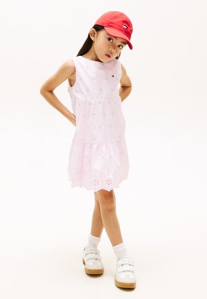 Junges Mädchen trägt ein hellrosa Kleid mit Blumen-Ausschnitten, rote Mütze, weiße Socken und weiße Turnschuhe, steht mit den Händen in der Hüfte.