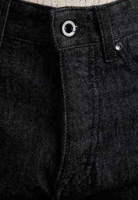 Mustat denim-farkut, joissa on tavallinen nappisuljenta, hopeiset yksityiskohdat ja hienovarainen tekstuuri. Näkyvät ompeleet saumoissa.